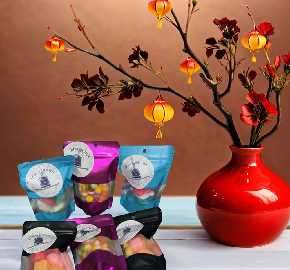 A table with assorted colorful packaged Crunchy Candy Treats in front of a red vase holding a decorative branch with flowers and hanging lanterns.